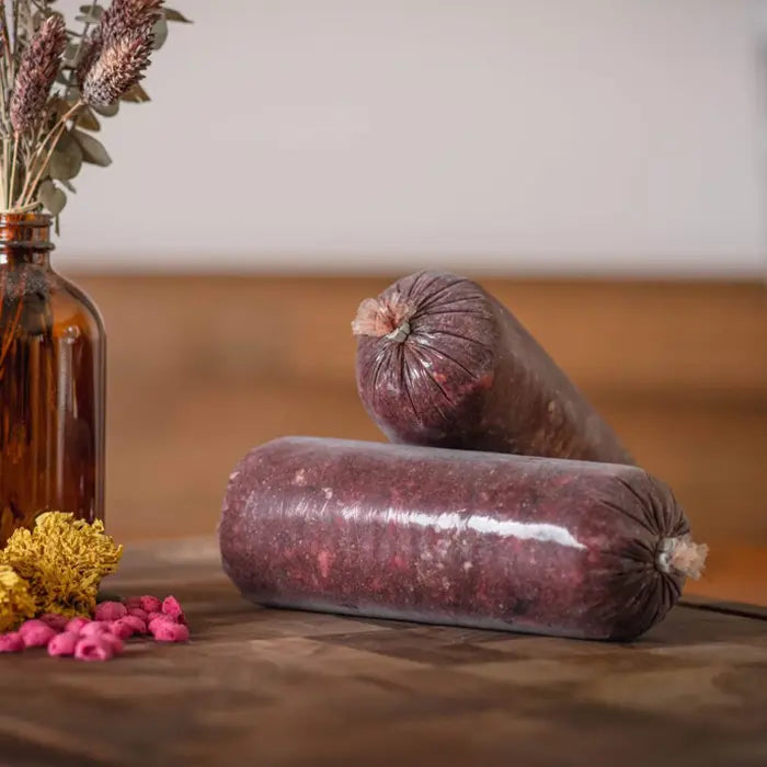 Two wrapped raw dog food tubes on a wooden surface with a vase of flowers in the background.